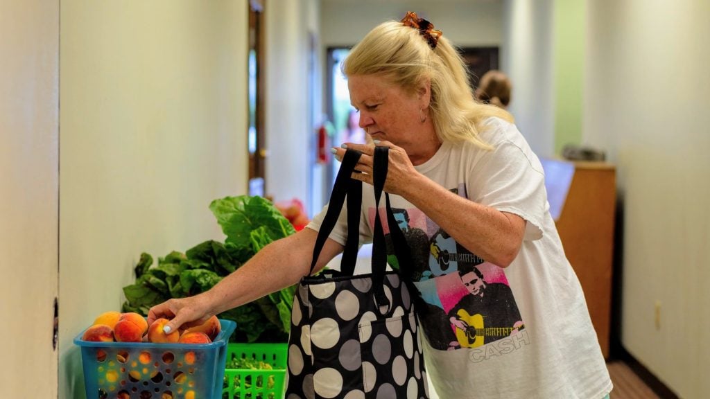 A woman holds a grocery bag in one hand and reaches for a peach with her other hand.