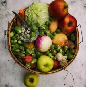 Example December Family Share A wicker basket filled with various fruits and vegetables, including apples, Brussels sprouts, carrots, onions, garlic, and a bag of seeds, on a light surface.