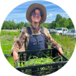 How-it-works_3 Farmer wearing blue overalls and a flannel carrying a black crate full of garlic scapes with a farm in the background