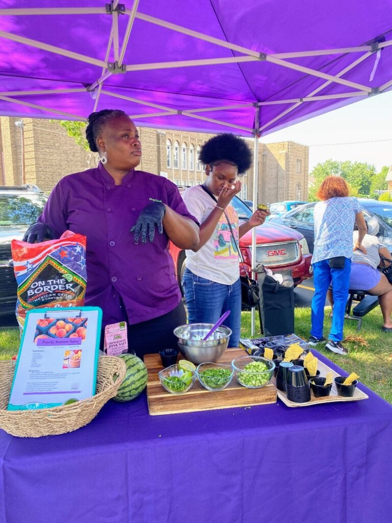 Chef wearing purple at an outdoor cooking demostration preparing a dish with yellow watermelon and tortilla chips